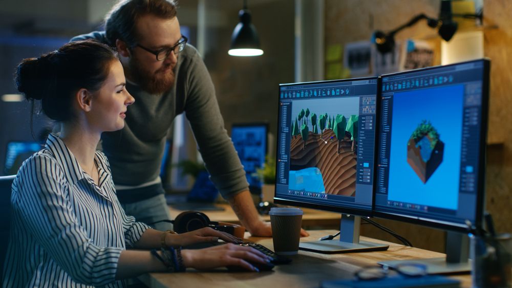 Female Game Developer Has Discussion with Male Project Manager While Working on a Game Level on Her Personal Computer with Two Displays.