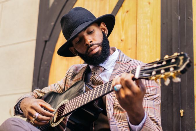 Stylish Black man in hat and classic outfit playing guitar outdoors