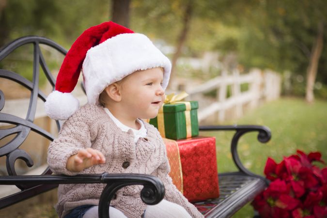 Young Child Wearing Santa Hat Sitting with Christmas Gifts Outside.