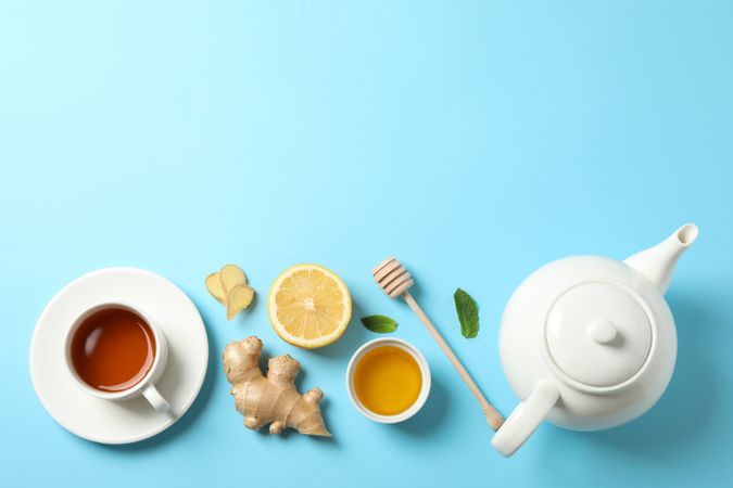Flat lay. Cup of tea, teapot, lemon, ginger, mint, honey and dipper on blue background, copy space