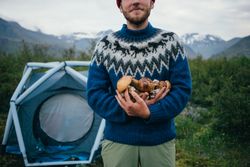 Man in front of tent wearing blue wool sweater cradling pile of foraged mushrooms