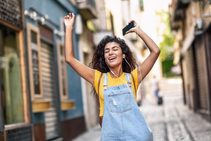 Arab woman listening to music with earphones and dancing in street with hands up