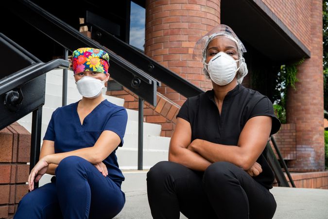 Two female nurses sit together on steps in front of hospital looking at camera