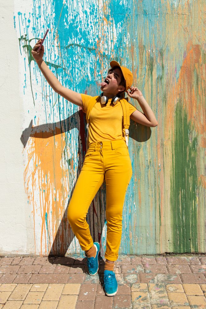 Trendy cool woman in yellow clothes taking selfie against painted wall in city