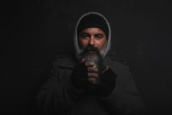 Homeless middle-aged man with a gray beard holding a cup of hot tea against dark background 5QXLV0