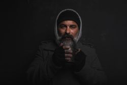 Homeless middle-aged man with a gray beard holding a cup of hot tea against dark background