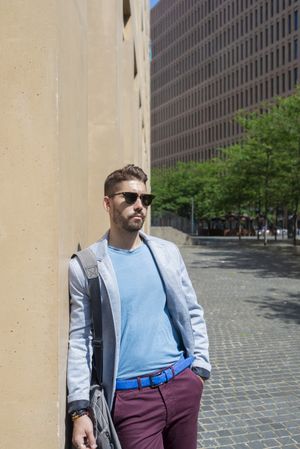 Bearded man in blazer leaning on a building wall outside with bag