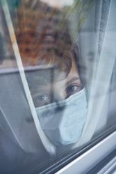 Cute boy in mask looking through window at home