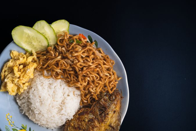 Flat lay of a plate with rice, fried fish, fried noodles, sliced omelette, and cucumber on a dark background
