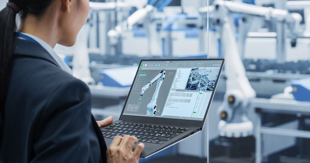 Manufacturing Robot Arms Administrator Programming Robotic Vision Systems to Execute Production at an Electronics Factory. Over the Shoulder Footage of a Female Supervising Assembly with a Computer