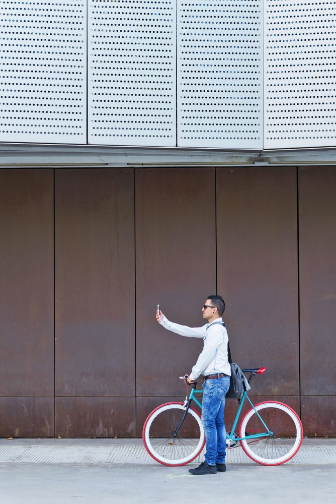 Male in sunglasses standing with red and green bicycle taking photo parked against a brown wall