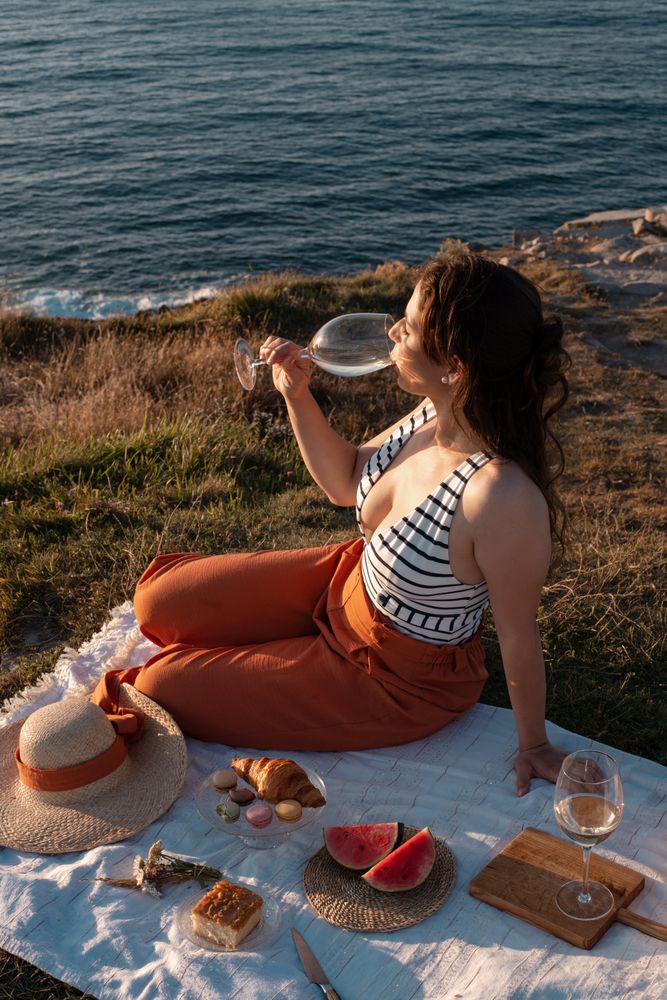 Woman with glass of drink on picnic mat looking at sea and mountains