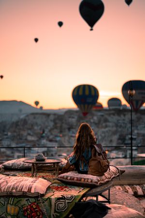Back view of woman sitting on cushions and watching the parachutes scene in Cappadocia, Central Anatolia, Turkey