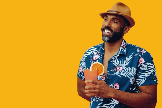 Smiling male holding a cocktail and looking away while wearing colorful shirt with yellow background
