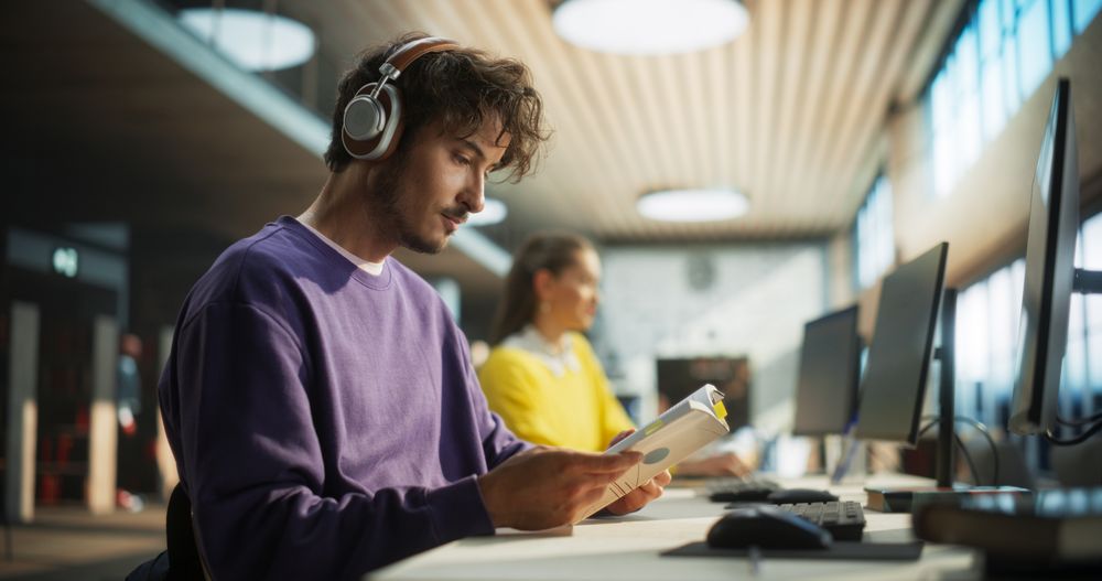 Handsome University Student Studying in a Traditional Public Library with Other Scholars. Young Male Wearing Headphones, Sitting in a Hall, Reading Academic Textbook and Preparing for Exams