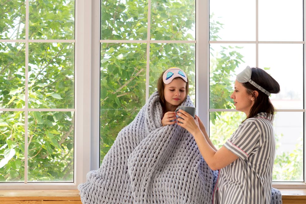 Mother comforting sick daughter by window with herbal tea.