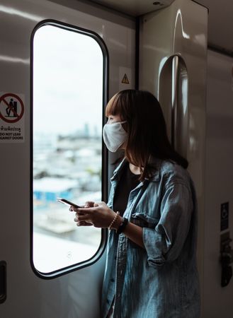 Woman with facemask standing in train looking through window holding smartphone