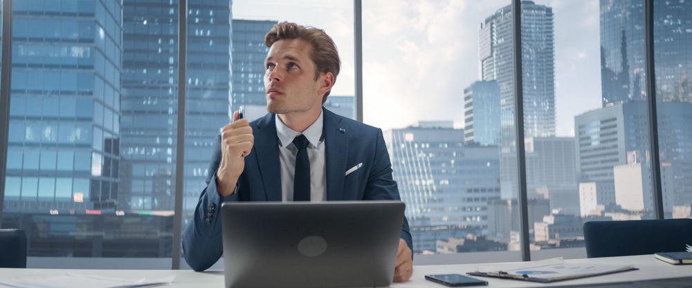 Confident Businessman in a Suit Sitting at a Desk in Modern Office, Using Laptop Computer