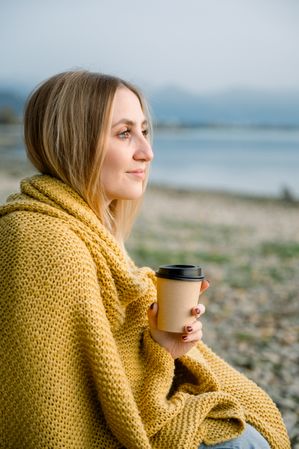 Young blond woman wrapped in blanket holding coffee mug at lakeshore