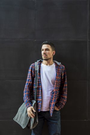 Man in plaid shirt standing outdoors with a bag on his shoulder on a sunny day