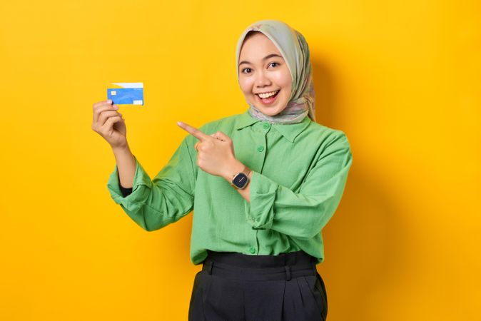 Muslim woman in headscarf and green blouse holding credit card smiling while pointing to it