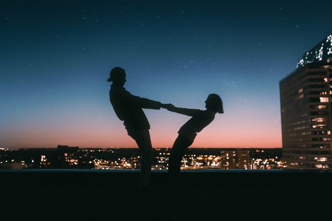 Silhouette of man and woman holding hands standing on rooftop near city at sunset