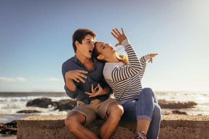 Couple making faces and hand gestures sitting near a sea shore