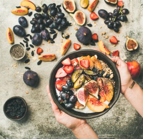 Woman and yogurt bowl over fruit assortment with figs, grapes, peaches, and chia seeds, square crop