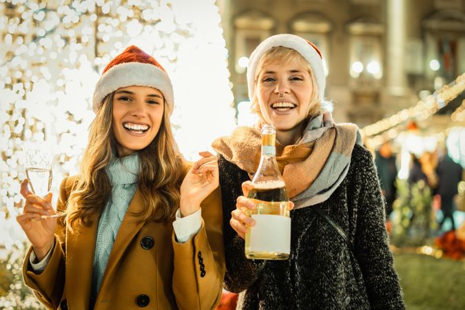 Two happy young women in Santa hats celebrating with champagne and looking at the camera.