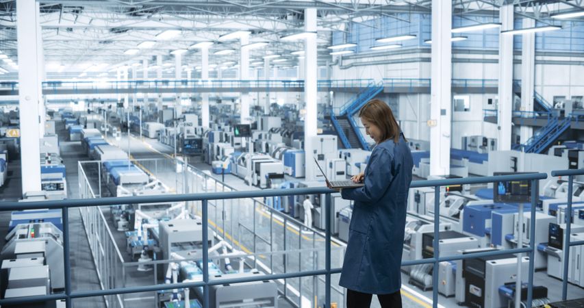 Young Asian Female Engineer Standing on a Platform, Using Laptop Computer at an Electronics Factory. Machines are Undergoing Maintenance, Specialist Monitoring the Progress Through Online Software