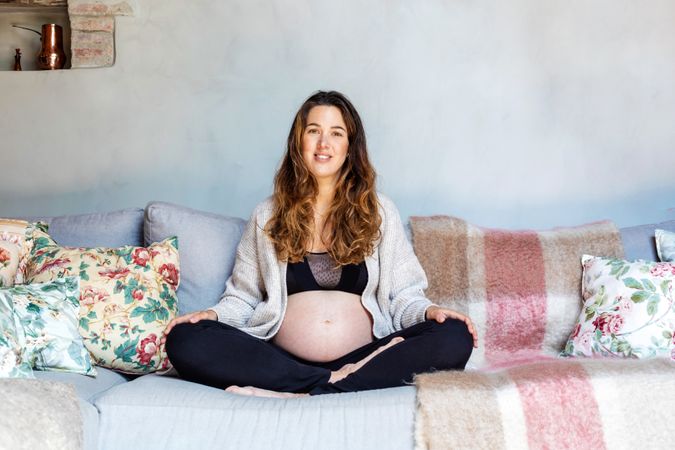 Pregnant smiling woman sitting cross legged on sofa