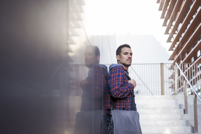 Man in plaid shirt leaning on wall outdoors in front of stairs on a sunny day