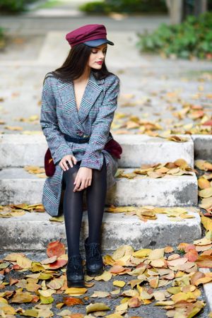 Female in warm winter clothes sitting among scattered fall leaves on park steps looking down