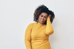 Black woman holding head in pain while wearing a yellow long sleeve shirt