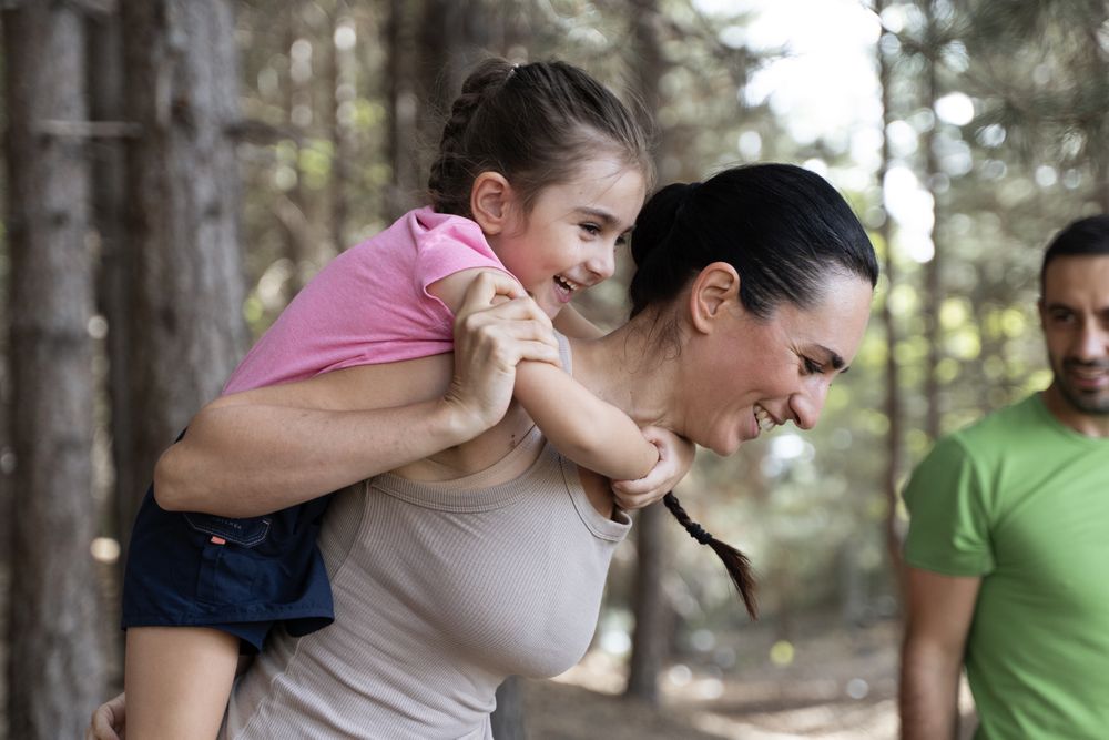 Happy mother giving her smiling daughter a piggyback ride, family having fun in the forest.