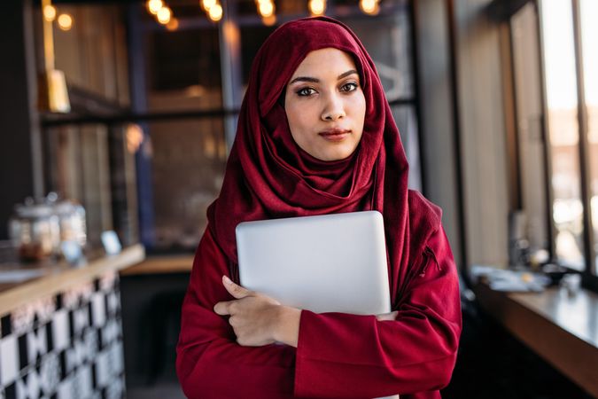 Confident woman in hijab with laptop standing indoors at a restaurant