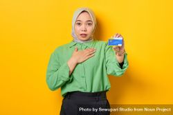 Muslim woman in headscarf and green blouse holding credit card with hand to her chest in surprise 486VY0