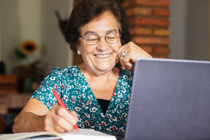 Older woman with glasses using a laptop and a notebook at home