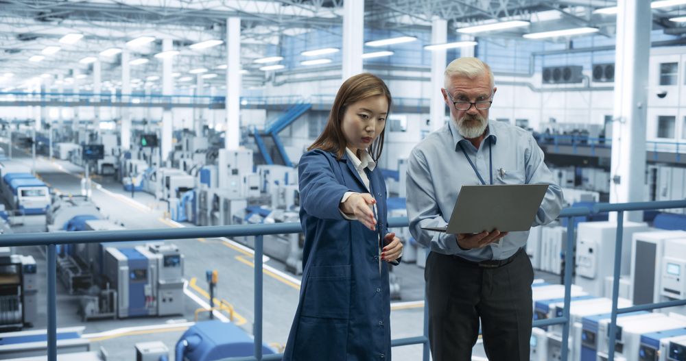 Mature Engineer and Young Japanese Technician Having a Conversation and Using Laptop Computer. Specialists Monitoring, Analyzing Software Reports at a Modern Electronics Factory