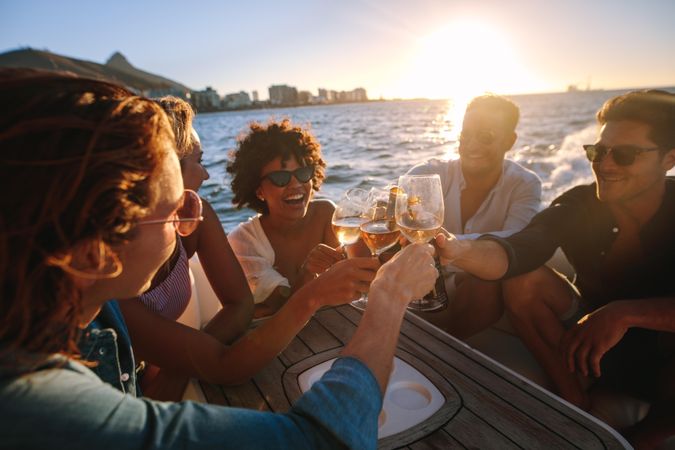 Cheerful young people on yacht drinking wine and beer with sun setting in bakground