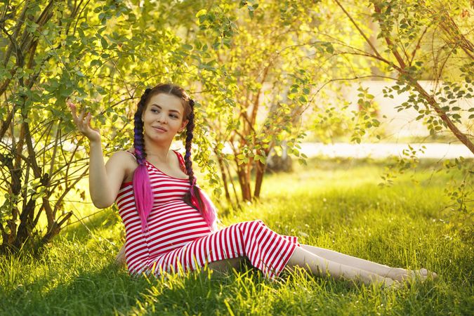 Pregnant female in red striped dress touching the trees while sitting on grass