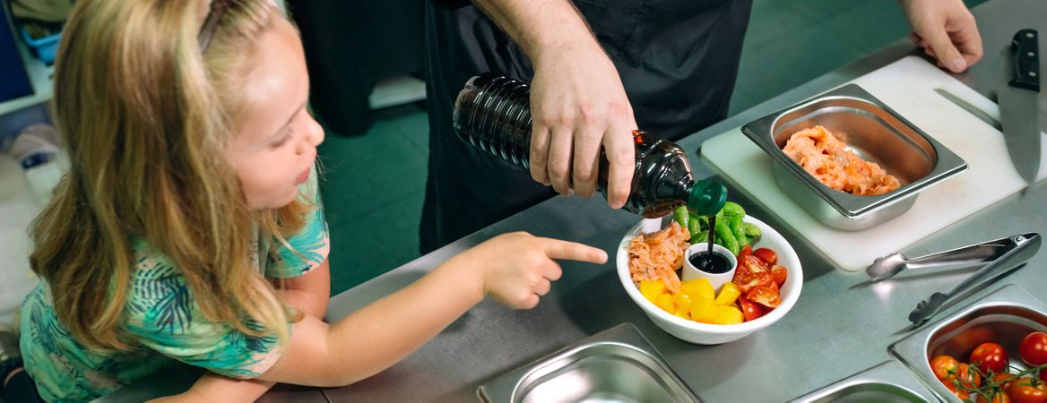 Banner of chef pouring soy sauce on poke bowl while happy girl pointing at it in restaurant kitchen