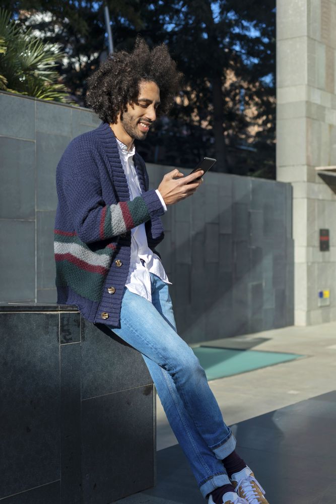 Smiling Black man using his smartphone while leaning on wall on sunny day