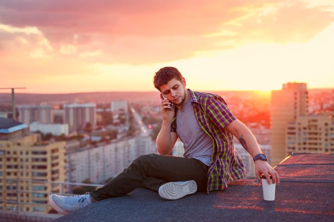 Male taking phone call while sitting on roof next to cup of coffee