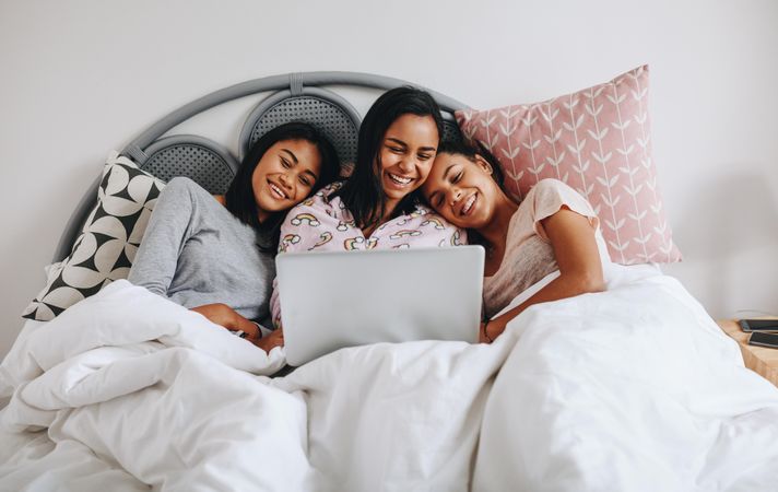 Smiling girls sitting on bed with laptop having fun at sleepover