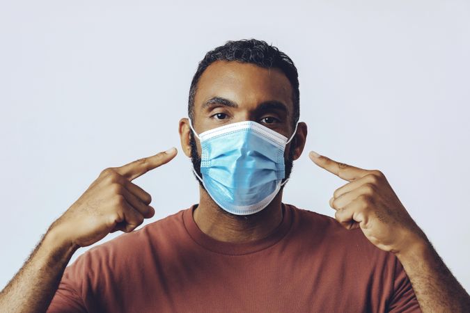 Male looking at camera while pointing at his face with a medical face mask on in studio shoot