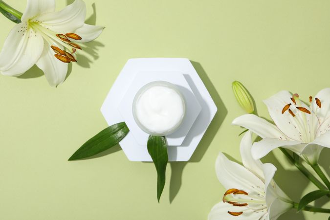 Jar with cream and lilies on green background, top view