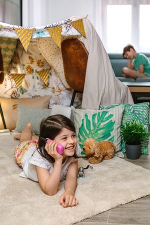 Smiling little girl holding walkie-talkie lying over rug in tent while talk with boy sitting on sofa