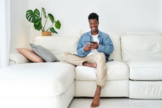 Cheerful Black man chatting on smartphone on sofa at home