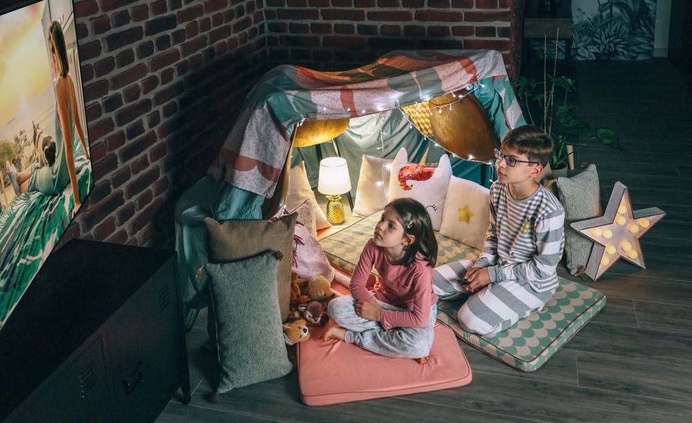 Boy and girl looking tv show sitting over mats on living room at night. Domestic life concept.
